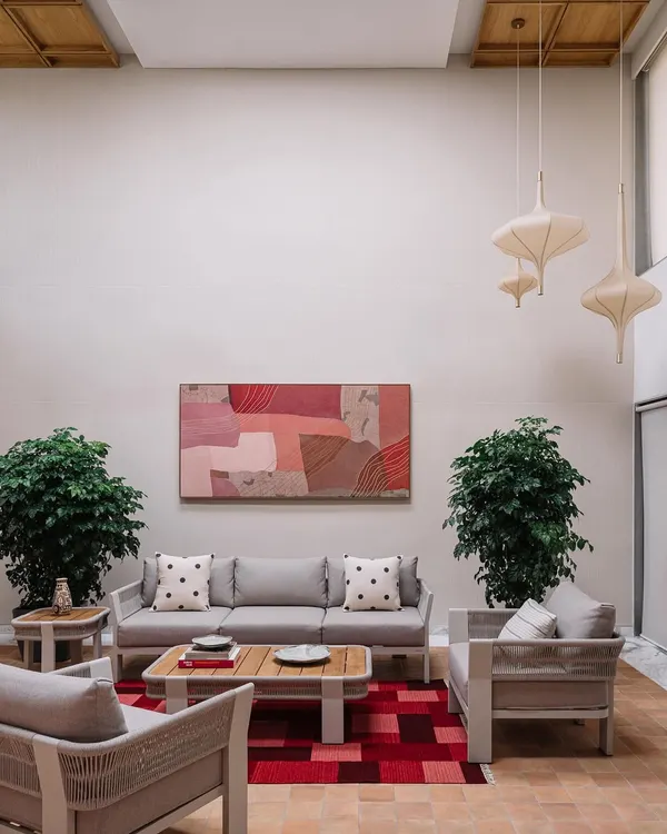 This living area in the Cocoon House is defined by its soaring double-height ceiling and a calm, neutral palette. We introduced warmth and personality with a large abstract painting in soft pinks, woven-texture furniture, and lush indoor plants, creating a space that feels both expansive and inviting.