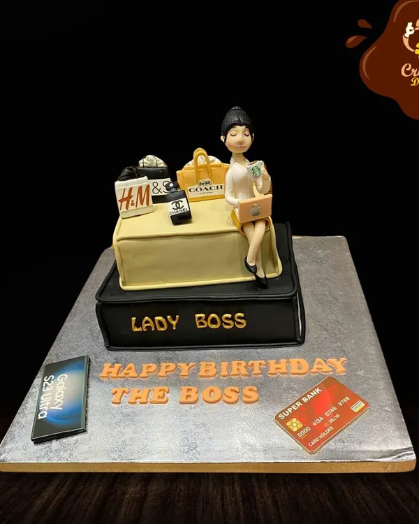 For the "Lady Boss" who does it all. This cake celebrates success with fondant figures of a boss lady, shopping bags from brands like H&M and Coach, a phone, and a credit card.
