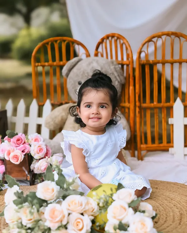 A close-up of a sweet baby girl during her first birthday photoshoot. Her gentle smile is captured perfectly in this outdoor setup, surrounded by soft pink flowers and a giant teddy bear.