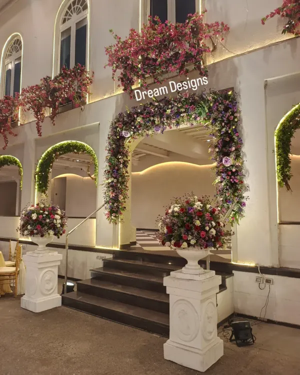 A beautiful floral entrance designed to complement the venue's architecture. The arch is covered in a rich tapestry of purple and red flowers, with matching arrangements in classic white urns.