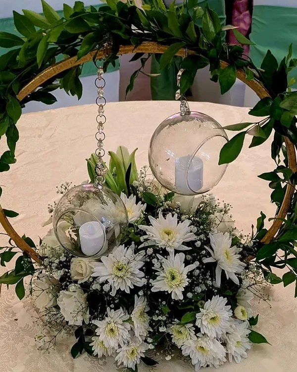 A close-up of an elegant table centerpiece. A hoop of fresh greenery encircles hanging glass orbs with candles, with a base of white chrysanthemums and baby's breath, creating a modern and romantic look.
