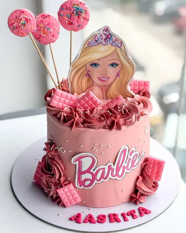 Come on Barbie, let's go party! This vibrant pink Barbie-themed cake is a dream for any fan. It's a buttercream cake decorated with a Barbie topper, pink chocolate bars, and matching cake pops, perfect for a fun and stylish birthday party.