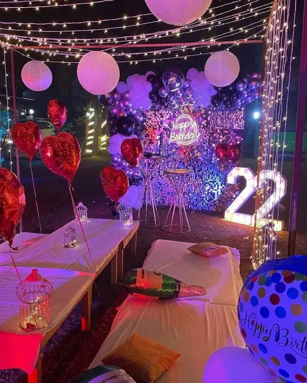 A beautiful outdoor cabana setup for a 22nd birthday. The space is decorated with fairy lights, heart-shaped foil balloons, and a statement backdrop with a neon sign and marquee numbers.