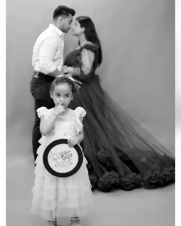 A creative family portrait where the older sister holds a sign announcing her promotion to "BIG Sister," while her parents share a kiss in the background.