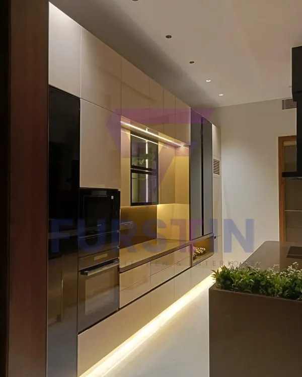 A perspective of the Bharatpur island kitchen, highlighting the seamless, handle-less drawers and the built-in wall oven. The warm, ambient lighting under the cabinets and along the floor creates a sophisticated glow, making the kitchen feel both modern and welcoming.