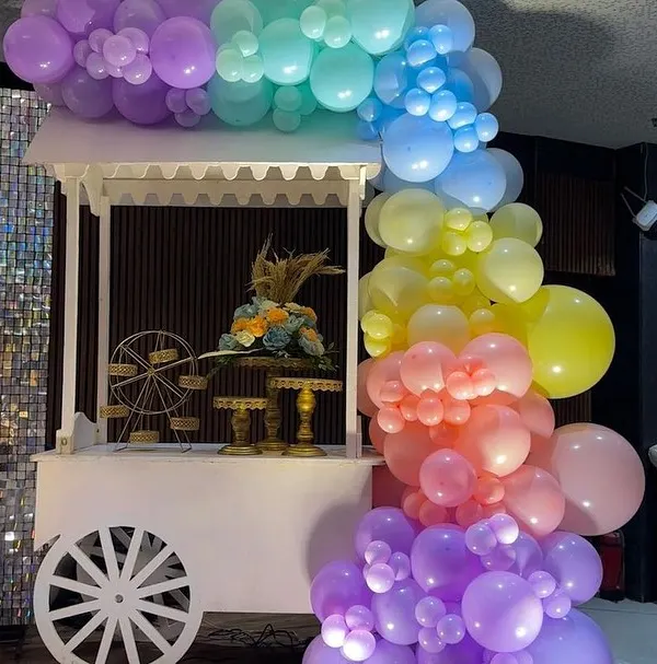 A charming white cart decorated with a pastel rainbow balloon garland, used as a dessert station for a beautiful baby shower.