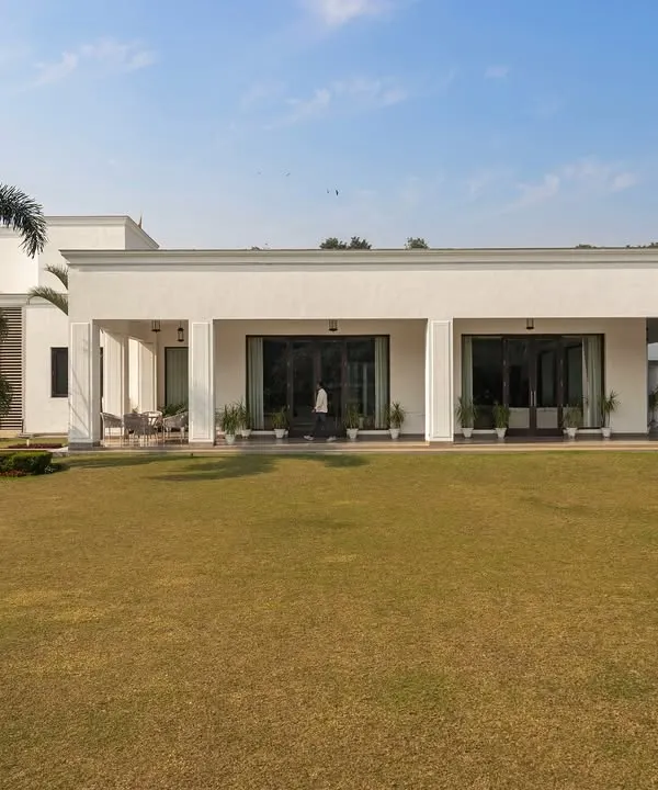 An exterior view of the Mehalin Farmhouse, where the landscape design emphasizes a vast, open lawn to accentuate the building's neo-classical form. The placement of palm trees frames the structure without obstructing the view.