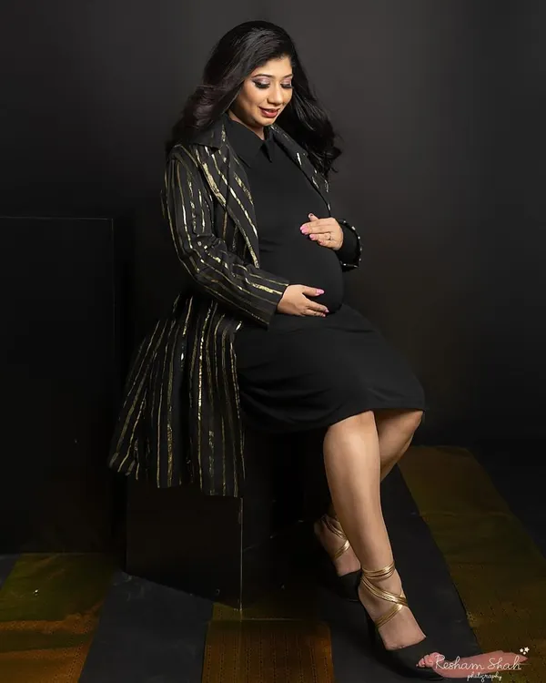 A touch of gold adds instant glamour. Here, the mom-to-be is seated, looking down at her bump, wearing a chic black dress paired with a sparkling gold-striped jacket for a sophisticated look.