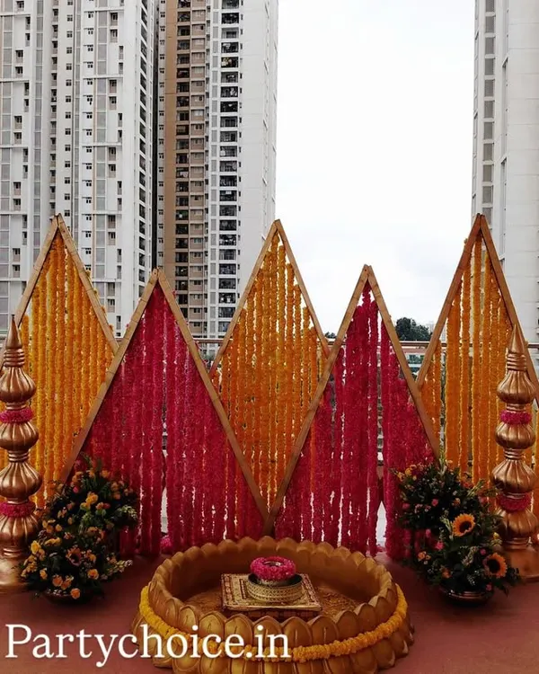 A striking backdrop for a Haldi or Harishinashasthra ceremony. The design uses geometric panels of yellow and pink marigolds, flanked by traditional golden props.