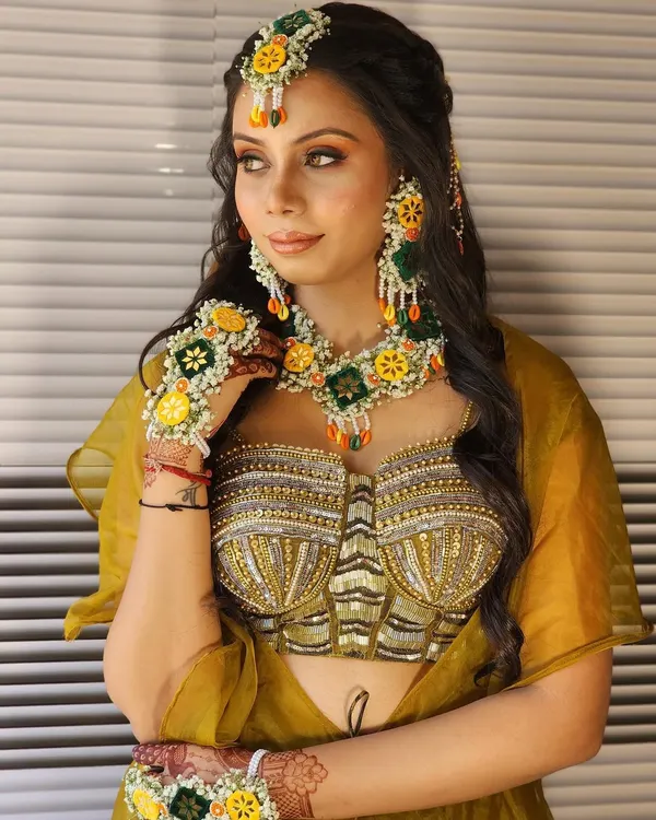 This is your dream mehendi look. The outfit features a stunning embroidered bustier in shades of gold and olive green, paired with floral jewelry for that perfect festive vibe.