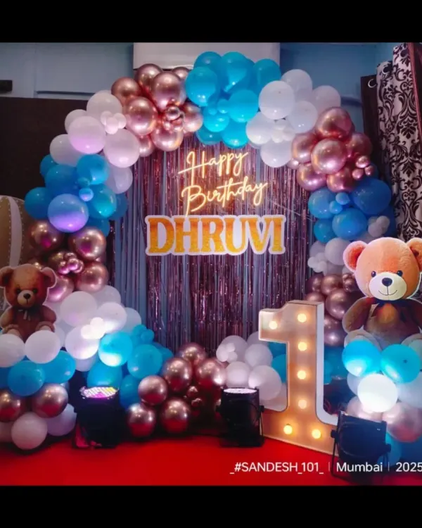 A beautiful circular balloon arch for Dhruvi's first birthday. The combination of rose gold, blue, and white balloons, along with cute teddy bears and a personalized neon sign, creates a warm and welcoming photo spot.