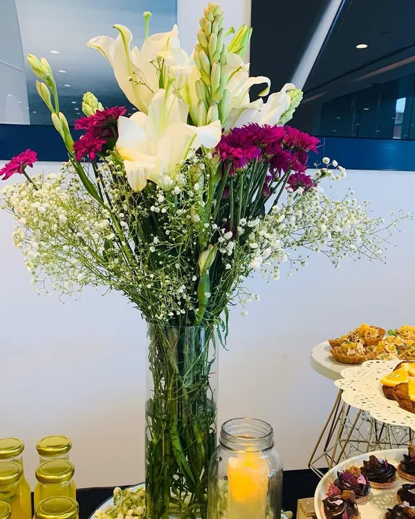 A classic and elegant floral centerpiece featuring white lilies, purple carnations, and baby's breath. Placed on a food and beverage station, arrangements like this add a touch of freshness and sophistication to any corporate event's catering setup.