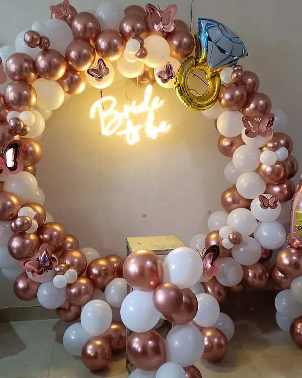 This is a complete bridal shower photo booth setup. It includes a balloon ring in rose gold and white, a "Bride to be" neon sign, and fun props like butterfly cutouts and foil balloons shaped like a diamond ring and a champagne bottle.