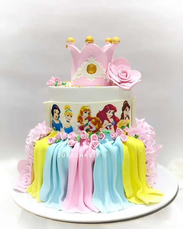 A grand three-tiered Disney princess cake featuring edible images of the princesses and beautiful fondant drapery. The design is topped with a handcrafted crown, making it fit for royalty.