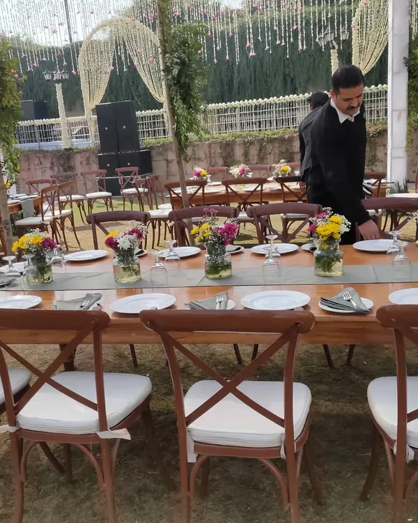 Long wooden tables paired with elegant cross-back chairs and padded cushions, creating a rustic-chic dining experience for an outdoor event.