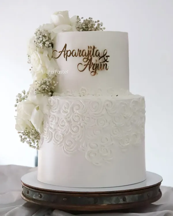An all-white, two-tier beauty. The bottom tier features a delicate hand-piped rosette pattern, while a cascade of white wafer paper flowers and baby's breath adds a touch of modern romance.