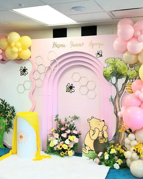 A sweet and charming Winnie the Pooh-themed setup for an Aqeeqah celebration, featuring a pink arched backdrop with honeycomb details. The scene includes a Pooh Bear cutout, a "hunny" tree, and a lovely balloon garland in pastel pink, yellow, and cream.
