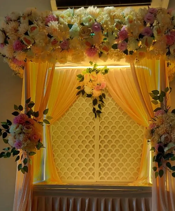 This decoration uses a warm, peachy color scheme with soft drapes and a thick arrangement of pastel-colored flowers. The integrated lighting from above and behind the lattice panel gives the entire setup a gentle, warm glow.