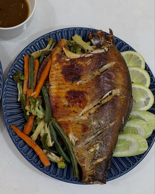 A whole roasted fish, marinated in our house Naga spices and roasted until the skin is crisp. It's served on a beautiful blue plate with steamed vegetables, fresh lemon, and a dipping sauce.