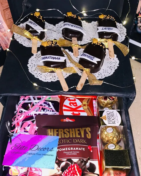The top layer of this surprise box reveals custom-made chocolate cakesicles with anniversary wishes. Below, a drawer is filled with an assortment of chocolates like Hershey's, KitKat, and Ferrero Rocher.