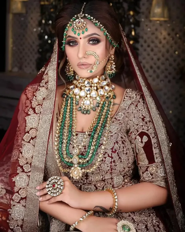 This is the ultimate royal bride look. The magnificent, multi-layered Kundan and emerald bead jewellery set is paired with a rich, embroidered maroon lehenga. It’s a timeless and grand statement for your wedding day.