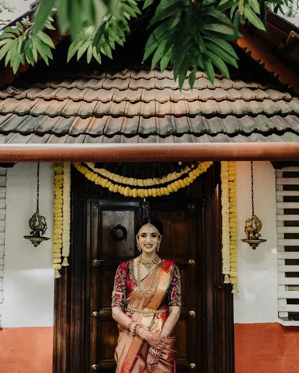 A beautiful full-length bridal portrait in front of a traditional wooden door. The symmetry and rich textures of the background perfectly complement the bride's elegant Kanjeevaram saree and jewelry.