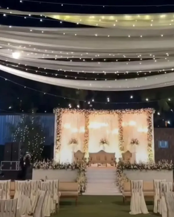 A wide-angle view of an elegant outdoor wedding reception, featuring a beautifully lit stage, guest seating with white covers, and a ceiling of fairy lights and drapes.