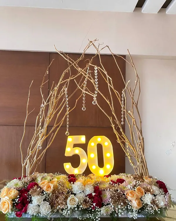 A four-foot-long floral centerpiece I created for a 50th birthday. It features light-up marquee numbers, an arch of gold-painted branches with hanging crystals, and a bed of roses and chrysanthemums.