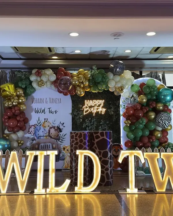 A full view of the "Wild Two" jungle safari stage we created for twins Vihaan and Vaniya. The setup includes a central balloon arch, a green foliage backdrop with a neon sign, and character cutouts, all framed by our popular light-up marquee letters.