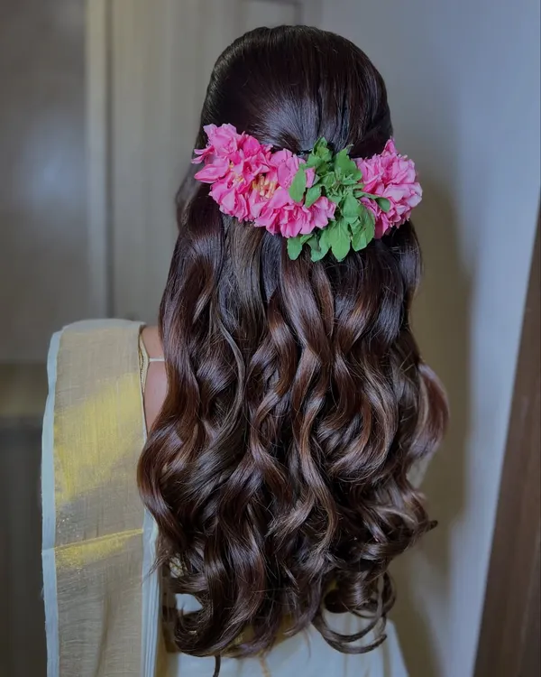 A beautiful half-up, half-down hairstyle with soft waves, adorned with a vibrant pink floral accessory for a South Indian bride.
