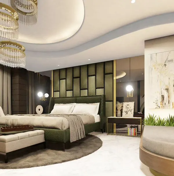 A luxurious master bedroom in a Noida penthouse, featuring a custom olive green upholstered headboard with geometric paneling. The room is designed with a curved false ceiling, elegant chandeliers, and a dedicated seating area for a suite-like feel.