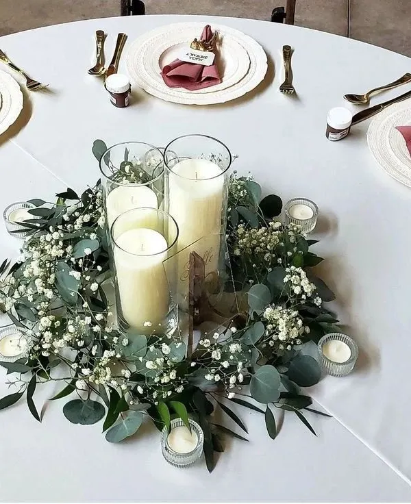A classic and elegant table centerpiece. The arrangement features white pillar candles in glass vases, surrounded by a wreath of baby's breath and eucalyptus leaves.