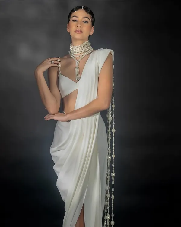 A studio shot for Nikhil Thampi's 'Gleam Collection', focusing on a modern white saree. The lighting is sculpted to highlight the intricate pearl jewelry and the unique drape of the garment against a dark, moody backdrop.