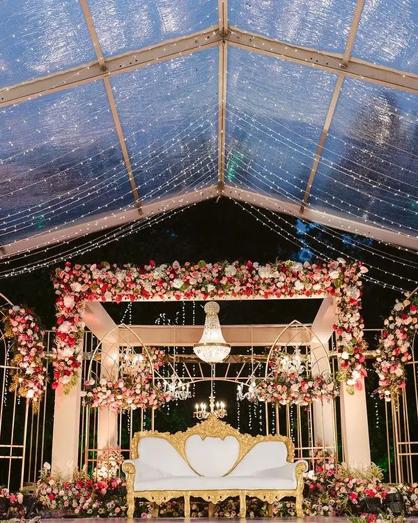 This contemporary reception stage is one of my favorites. I combined elegant gold-arched frames with lush floral arrangements and sparkling chandeliers under a clear tent, creating a magical, starlit atmosphere for the couple's first celebration.