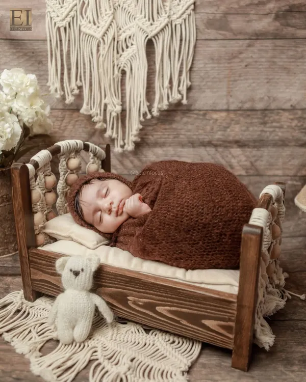 A baby swaddled in a warm brown wrap, sleeping peacefully on a rustic wooden bed with macrame details. This setup showcases our love for natural textures and tones.