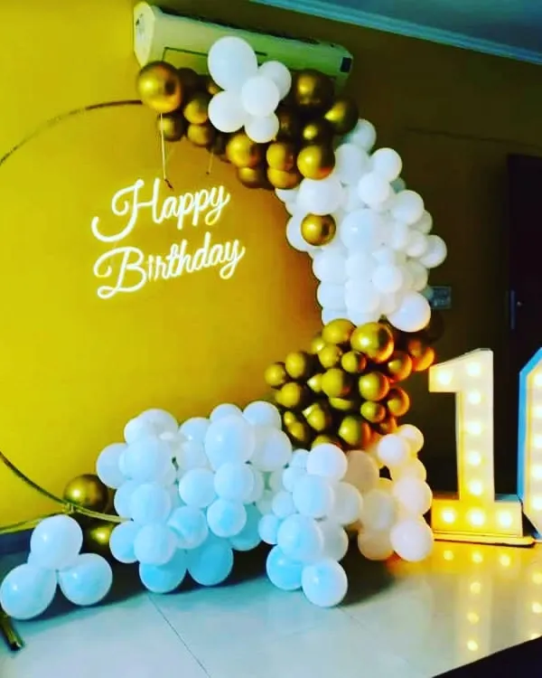A chic 16th birthday setup with a gold and white balloon arch on a ring frame. The large light-up '16' numbers and 'Happy Birthday' neon sign make it a perfect spot for birthday photos.