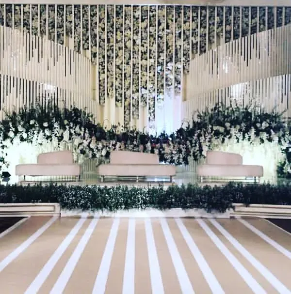 A modern and sophisticated reception stage design featuring clean lines and a neutral color palette. The backdrop uses white floral arrangements and textured panels to create a chic, minimalist look for a contemporary wedding.