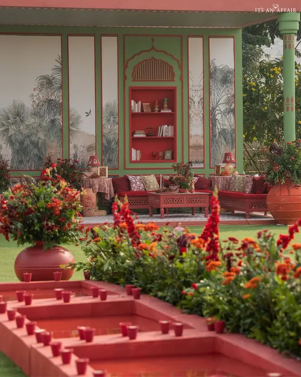 A lounge area from 'A Mughal Garden' wedding, featuring rich red sofas against a sage green wall with hand-painted murals. The design creates a luxurious, library-like feel in an outdoor setting.