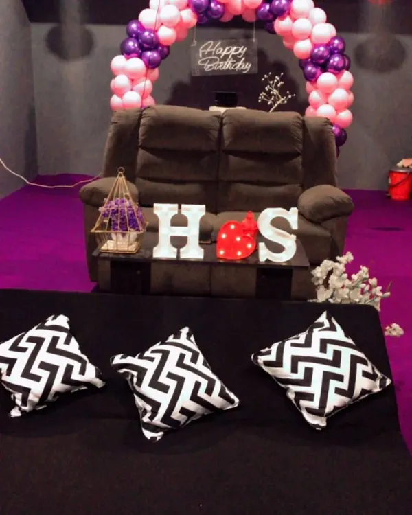 A cozy birthday setup perfect for a couple or a few friends. This room features a pink and purple balloon arch, a comfortable recliner, and custom initial lights, creating an intimate space for your private birthday movie screening.