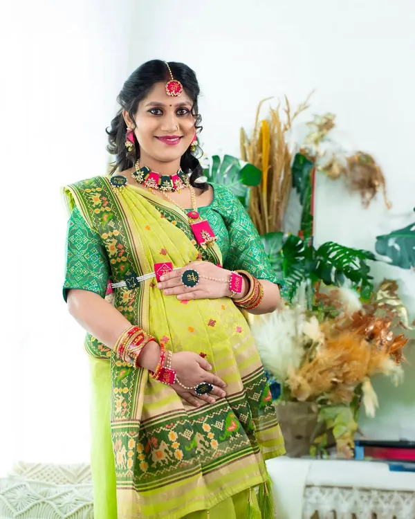 A vibrant and traditional portrait. The mom-to-be is dressed in a beautiful green saree with intricate details and matching floral jewelry, celebrating her pregnancy with cultural pride.