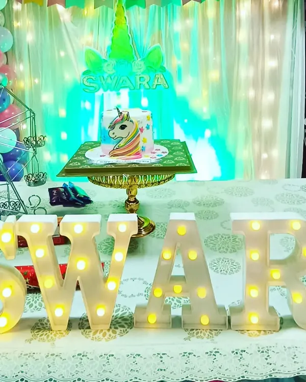 A magical unicorn theme for Swara's birthday. The setup features a beautiful cake table with a unicorn cake, marquee letters of her name, and a backdrop with fairy lights, creating a whimsical photo corner.