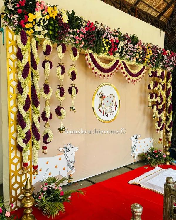 A beautiful and auspicious cow-themed backdrop for a traditional ceremony. The setup features hanging floral arrangements and illustrations of Kamadhenu, the sacred cow, symbolizing purity and prosperity.