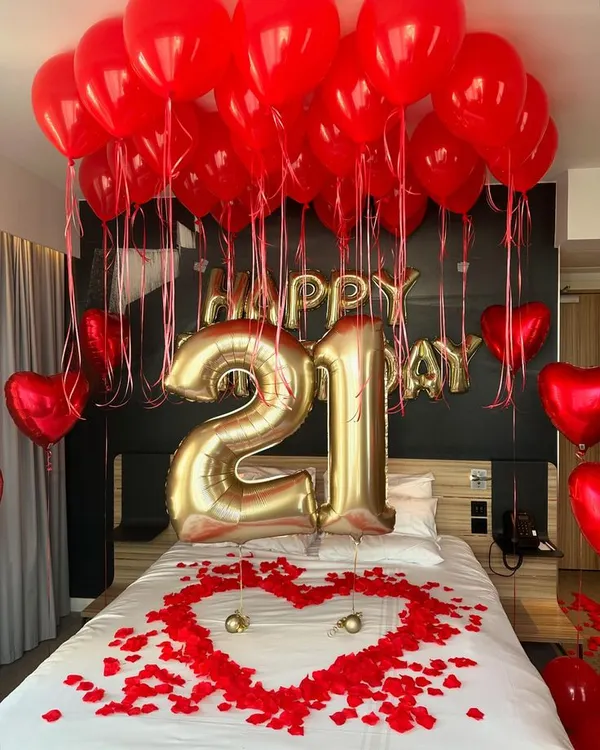 A classic romantic birthday surprise featuring a bold red and gold color scheme. The decoration includes large "21" foil balloons, "Happy Birthday" lettering, and a heart made of rose petals on the bed.