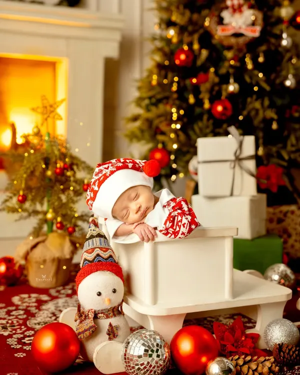 A perfect Christmas dream. This newborn sleeps peacefully in a festive setup with a warm fireplace, a decorated tree, and shiny ornaments.