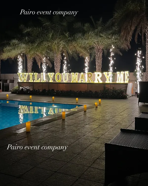 This poolside proposal setup features large, illuminated "WILL YOU MARRY ME" letters and dramatic cold pyro sparklers that light up the night. The palm trees and glowing candles along the pool add to the romantic resort-like atmosphere.