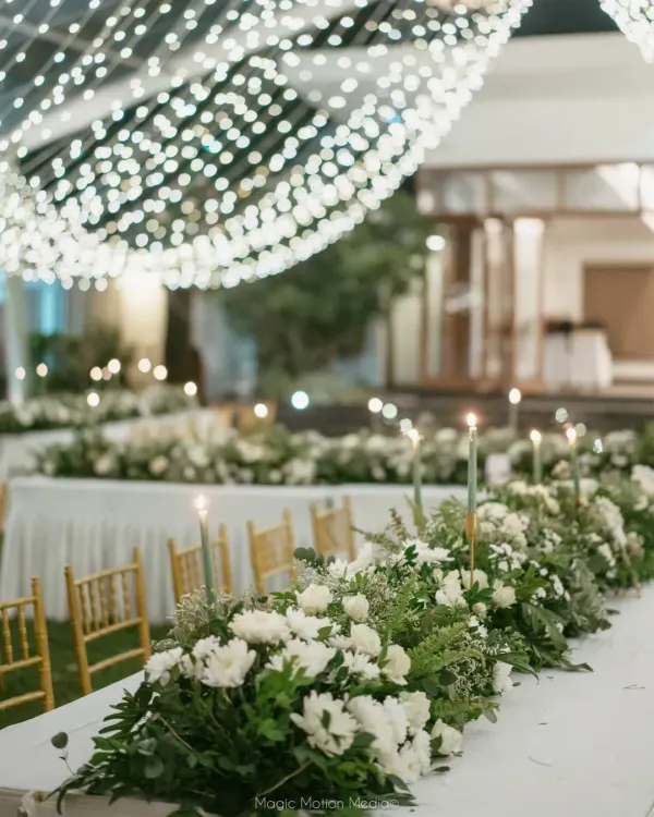 A vision in white. For this elegant cocktail dinner, we designed a stunning tablescape with a lush runner of white flowers and greenery, set under a transparent roof filled with fairy lights for a magical, starlit dining experience.