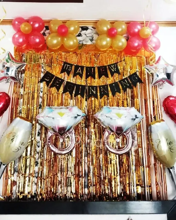 This engagement party decoration is perfect for celebrating a new beginning. The setup includes a gold tinsel curtain, foil balloons shaped like diamond rings and champagne glasses, and a celebratory banner.