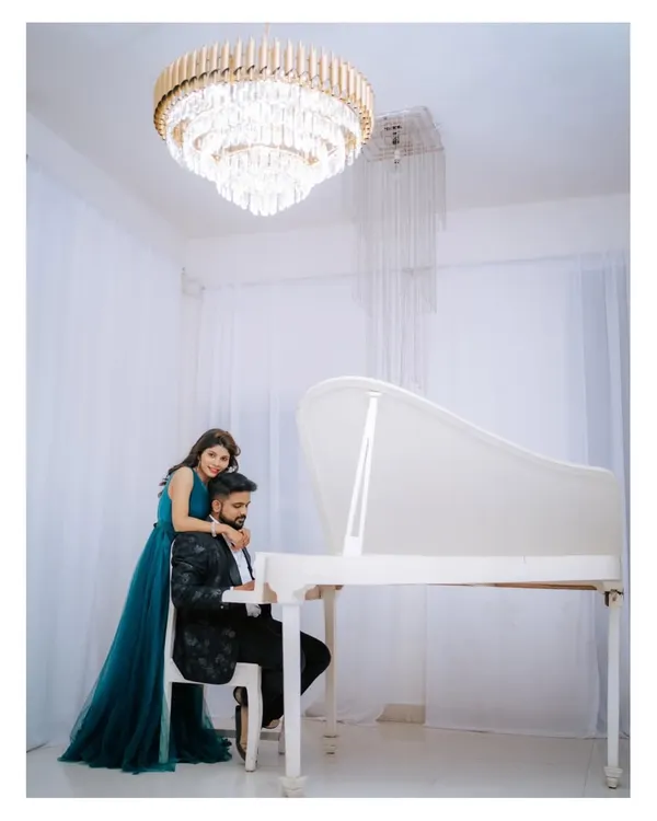 An elegant pre-wedding portrait of a couple in western attire, posing by a grand white piano under a magnificent crystal chandelier.