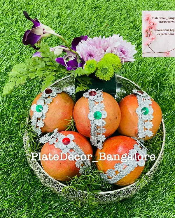 Pomegranates decorated with silver lace and kundan stones, arranged in a silver filigree bowl with fresh flowers. This is a classic and elegant fruit platter design.