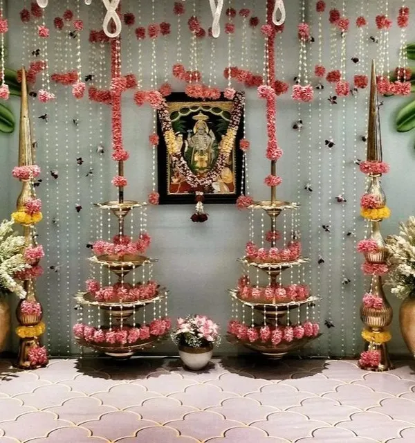 An elegant decor setup for a bridal pooja at home. The design uses traditional brass lamps (deepams), strings of jasmine and pink flowers, and a framed picture of a deity to create a serene and sacred space.